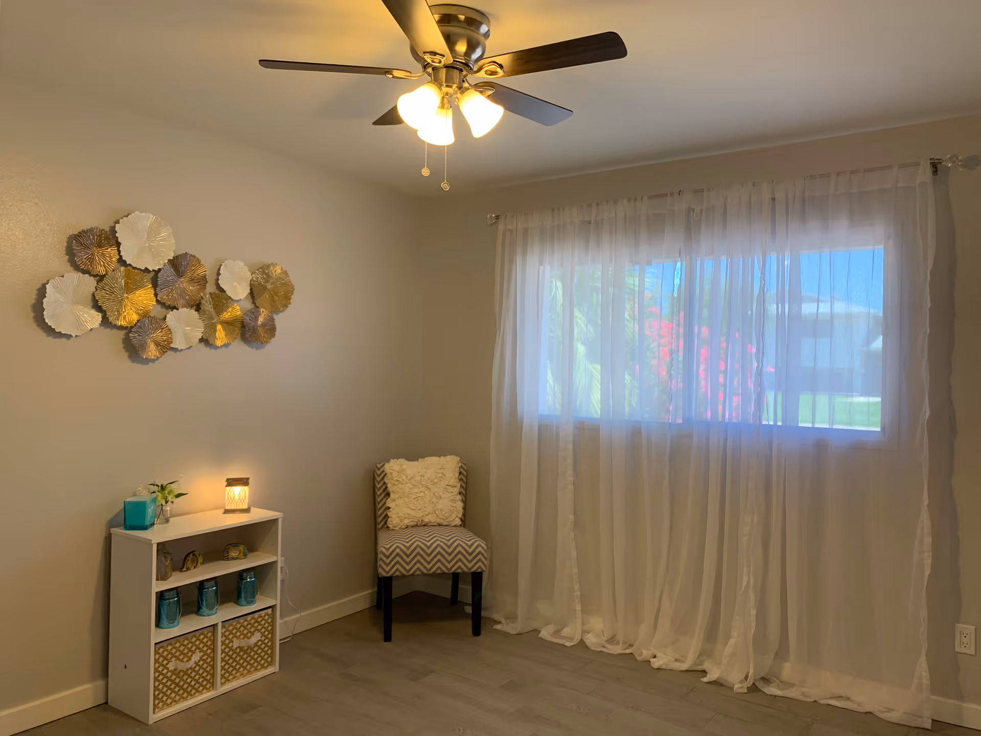 A cozy room with a ceiling fan and light fixture, a window covered with sheer white curtains, a small white shelf with decorative items and a lit candle, a patterned chair with a white cushion, and a wall decoration made of metallic circular shapes.