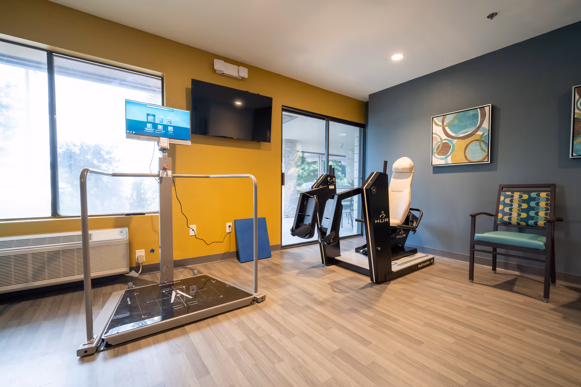 A small exercise room with light wood flooring and walls painted in mustard yellow and dark gray. The room contains a balance platform with handrails and a mounted screen, a seated exercise machine, a patterned chair, and two abstract paintings on the wall. Large windows and a glass door provide natural light and a view of an outdoor patio area.