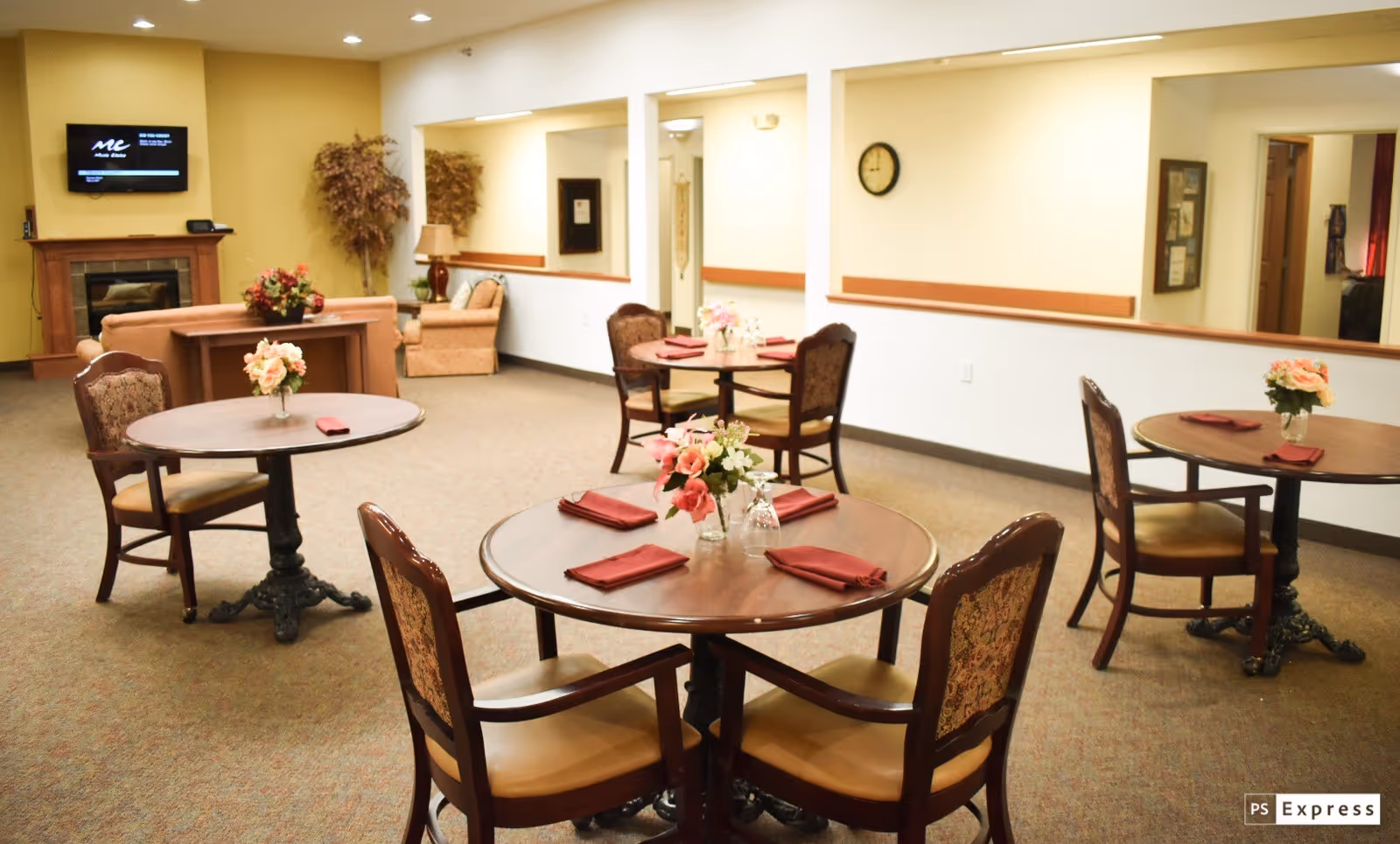 A senior living facility dining area with round wooden tables, each set with red napkins and floral centerpieces. The room has beige walls, carpeted floors, and a cozy seating area with armchairs near a fireplace. A wall-mounted TV is visible above the fireplace.