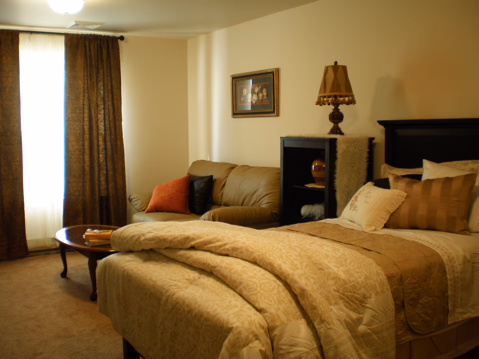 A cozy bedroom with a neatly made bed featuring beige and brown bedding and multiple pillows. Next to the bed is a small black shelving unit with a decorative lamp and vase. A beige sofa with orange and black cushions is positioned near a window with brown curtains. A wooden coffee table with a book and glasses sits in front of the sofa.