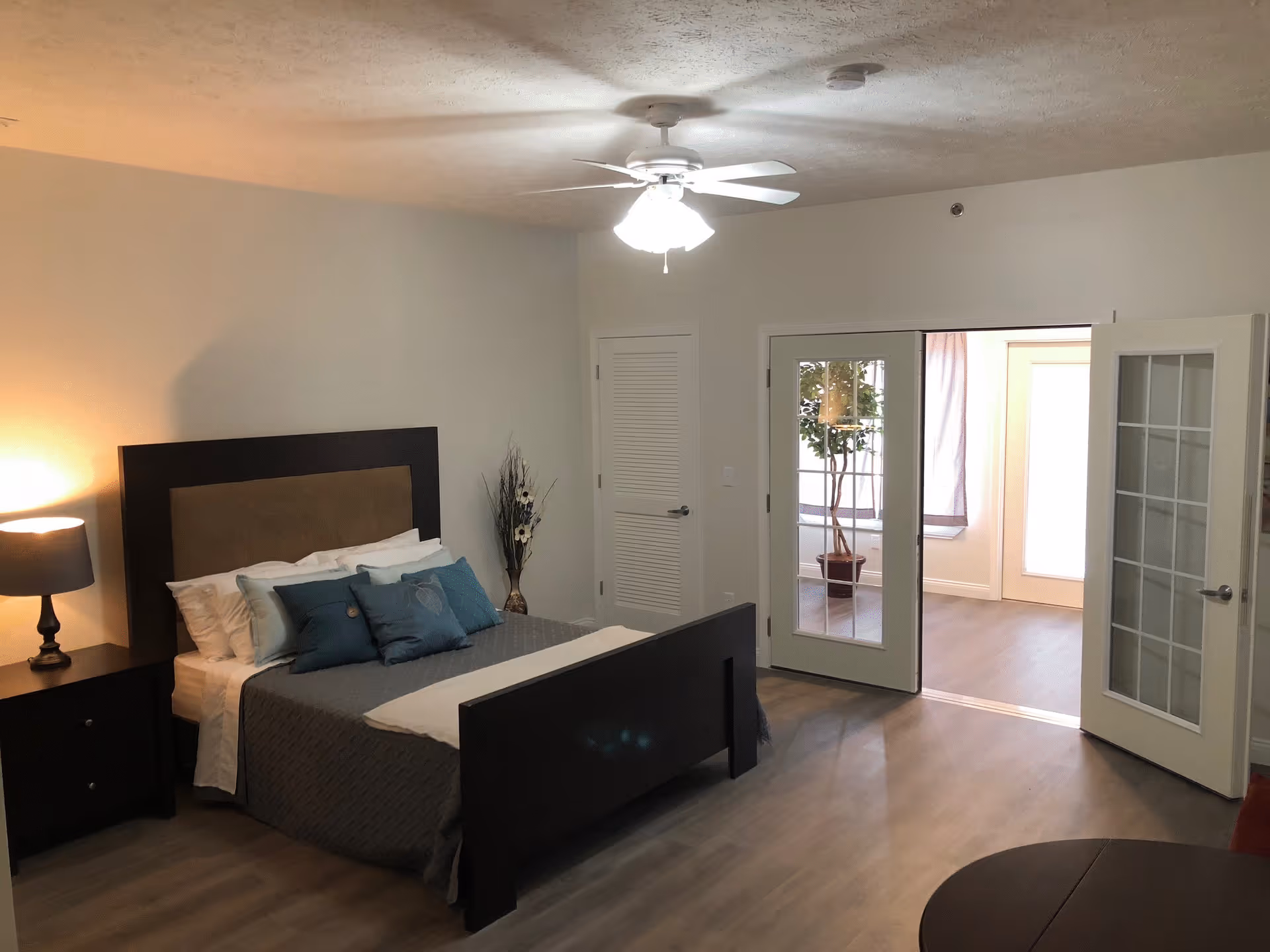 A bedroom with a dark wooden bed frame and headboard, neatly made with white and blue pillows and a gray quilt. To the left of the bed is a nightstand with a lamp. The room has light-colored walls and wood flooring. There is a ceiling fan with a light fixture above. French doors open to an adjacent room with a potted plant and large windows.