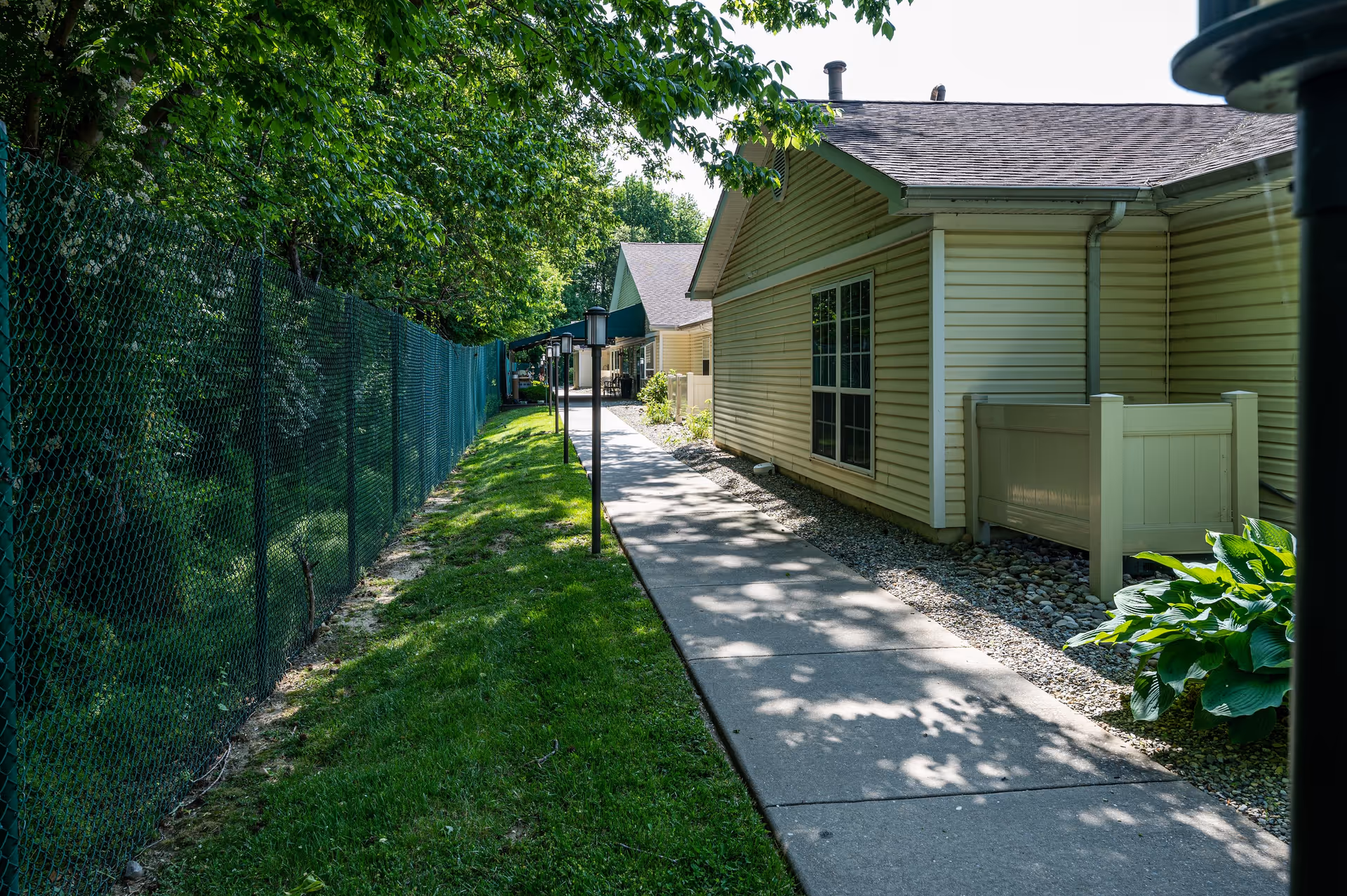 A paved walkway runs alongside a beige building with a window, bordered by a green lawn and a chain-link fence on the left. Trees provide shade over the path, and there are lamp posts along the walkway.