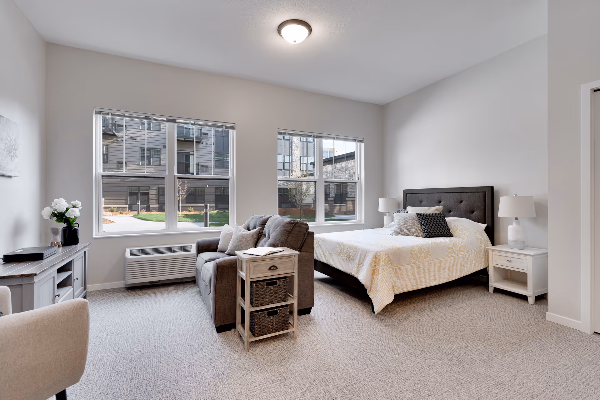 A bright and spacious bedroom with a large bed featuring a dark upholstered headboard and white bedding with decorative pillows. Next to the bed are two white nightstands with matching white lamps. In front of the bed is a gray loveseat with a side table holding a book. The room has two large windows letting in natural light and showing an exterior view of a courtyard and neighboring buildings. The walls are painted light gray and the floor is carpeted in a neutral tone.