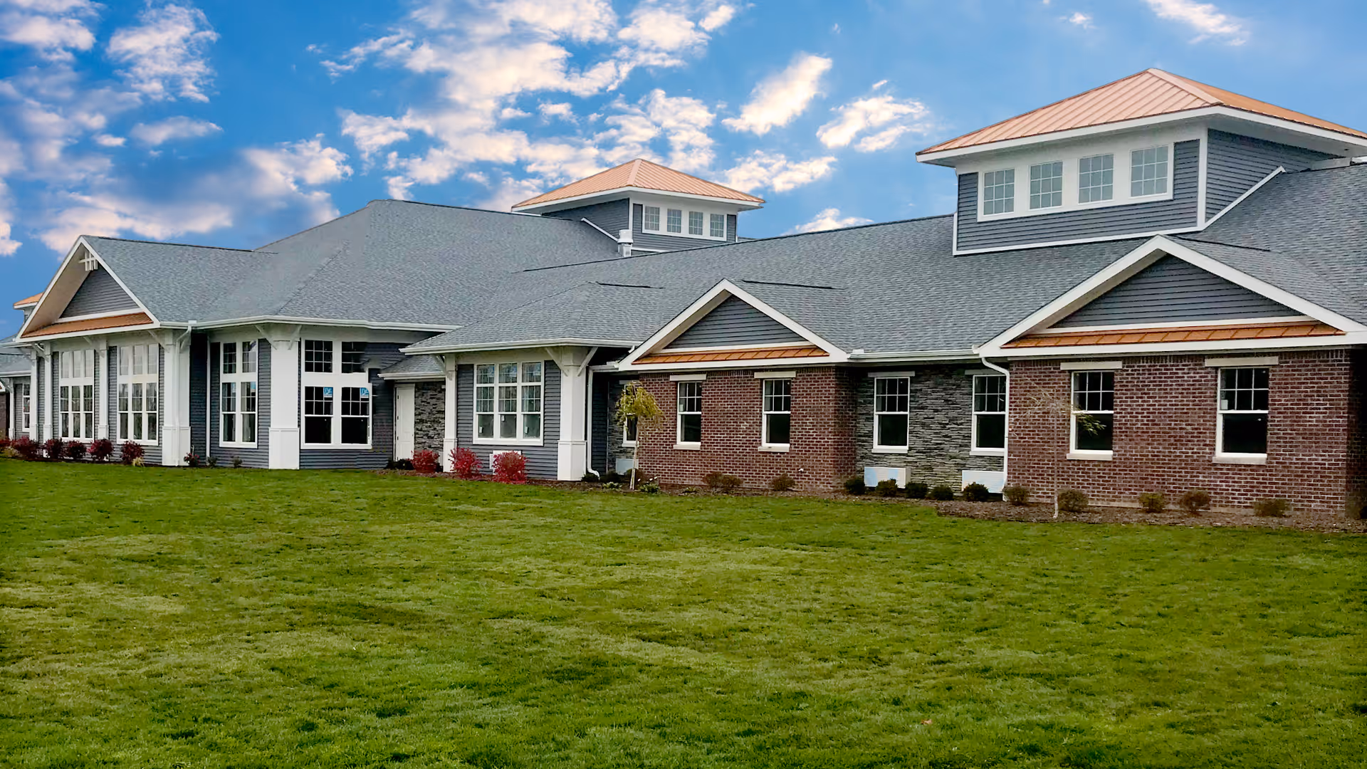 Exterior view of a single-story senior living facility building with a combination of brick and siding walls, multiple windows, and a well-maintained green lawn under a partly cloudy blue sky.
