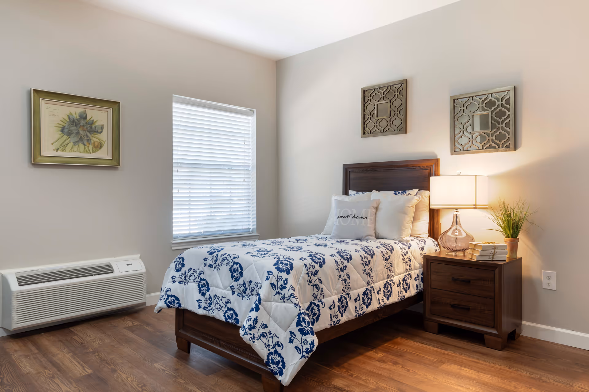A cozy bedroom with a single wooden bed featuring a white and blue floral comforter and several pillows, including one with the words 'sweet home'. Next to the bed is a wooden nightstand with a glass lamp, a small plant, and a stack of books. The room has light-colored walls, a window with white blinds, a framed botanical print on one wall, and two decorative mirrors above the bed. The floor is wooden.