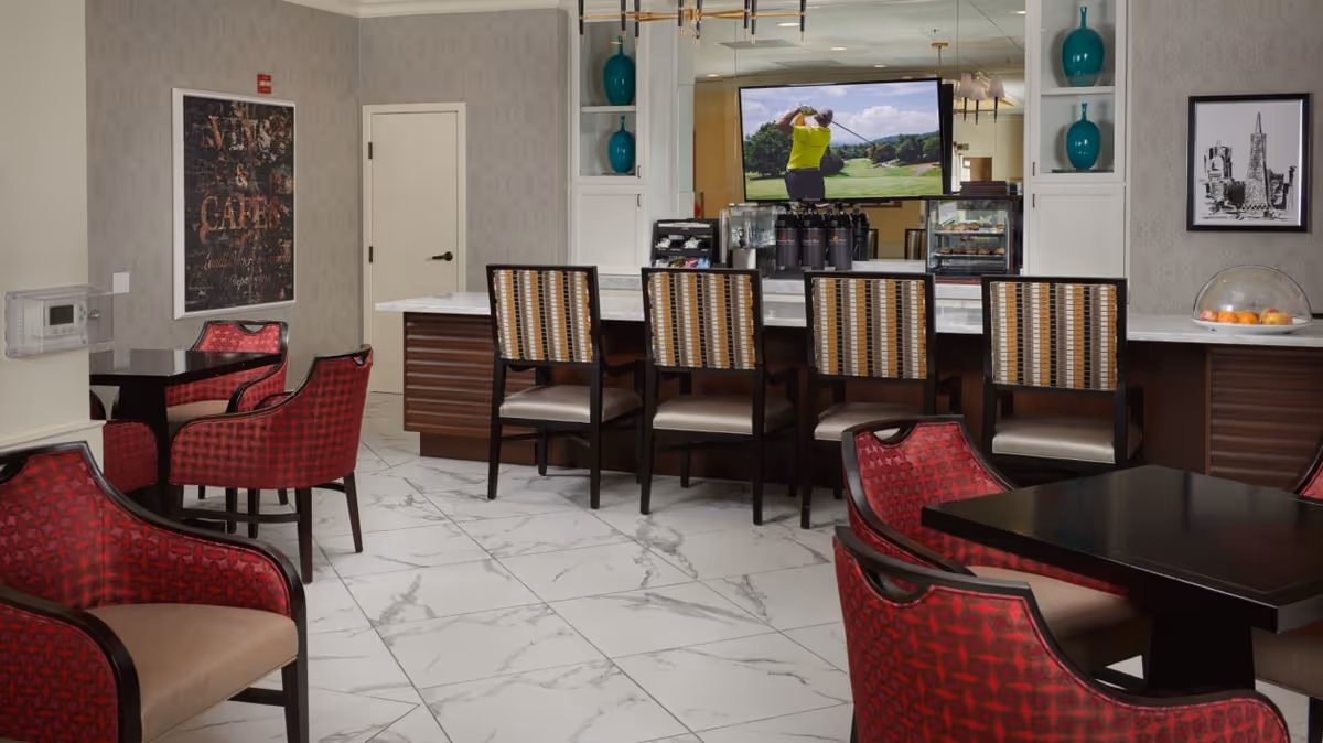 A modern dining area with a marble floor, featuring a counter with four striped chairs and a television mounted above it showing a golfer in mid-swing. There are red cushioned chairs around dark wooden tables in the foreground, decorative teal vases on shelves, and framed artwork on the walls.