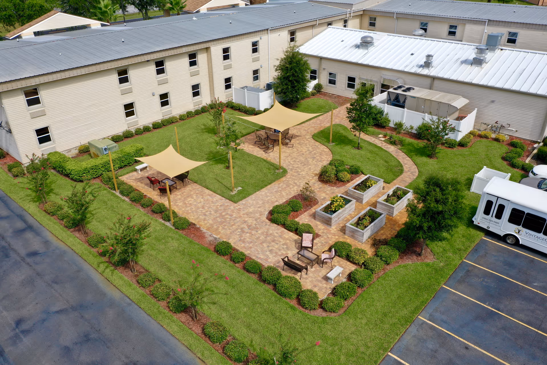 Aerial view of a landscaped outdoor courtyard area at a senior living facility with paved walkways, shaded seating areas with tables and chairs, raised garden beds, and surrounding greenery. The beige building with multiple windows encloses the courtyard, and a white shuttle bus is parked nearby in the parking lot.