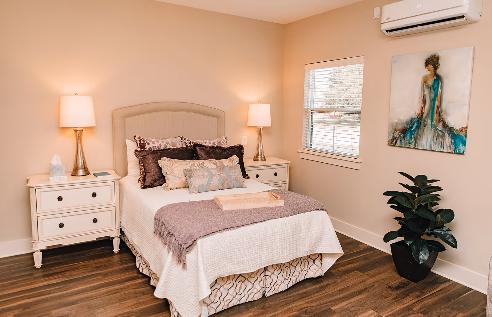 A cozy bedroom with a neatly made bed featuring multiple decorative pillows and a purple throw blanket. There are two white nightstands on either side of the bed, each with a lamp. A window with blinds is on the right wall, and a potted plant sits below a colorful painting of a woman in a blue dress. The room has wooden flooring and beige walls.