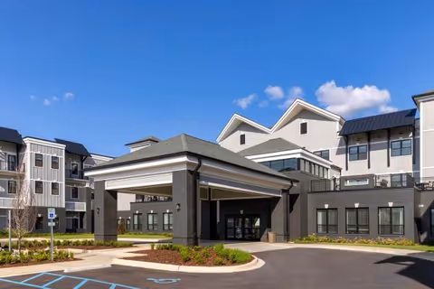 Exterior view of a modern senior living facility building with a covered entrance, multiple windows, and a clear blue sky above. The driveway and parking area in front include marked handicap parking spaces and landscaped greenery.