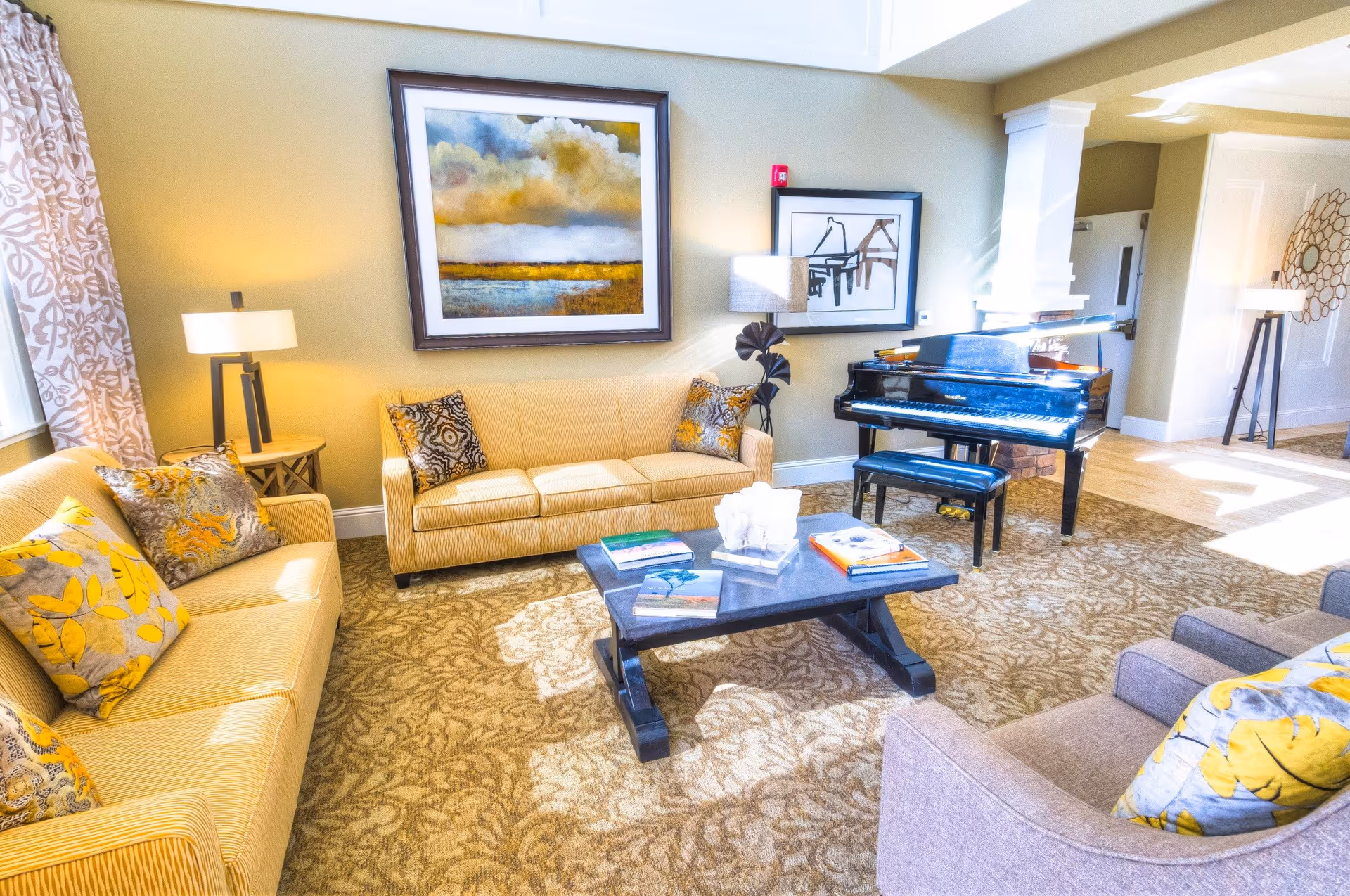 A bright and cozy living room with two yellow sofas and a gray armchair, all adorned with patterned yellow and gray pillows. A black grand piano with a matching bench is placed against the wall. The room features a patterned carpet, two framed artworks on the walls, and two floor lamps providing warm lighting. Sunlight streams in from a window with patterned curtains on the left side.