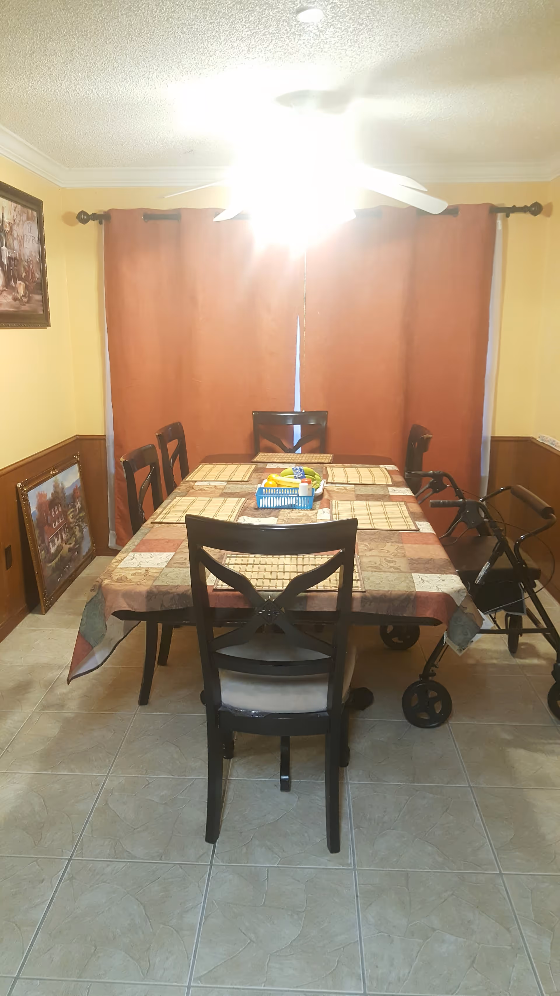 A dining room with a rectangular table covered with a patterned tablecloth and six chairs around it. There is a basket with fruit and other items on the table. The room has tiled flooring, yellow walls with wooden paneling, and a ceiling fan with a bright light. A walker is positioned next to one of the chairs, and two framed pictures are visible, one hanging on the wall and one leaning against the wall.