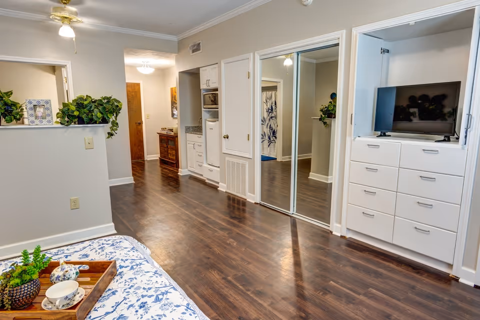 Interior view of a senior living facility room with wooden flooring, white walls, and ceiling fans. There is a bed with blue and white floral bedding partially visible in the foreground with a wooden tray holding a teapot, cup, and small plant. On the right side, there is a built-in white dresser with a flat-screen TV on top and mirrored sliding closet doors. The hallway leads to other rooms with a wooden door visible at the end and some green plants decorating the space.