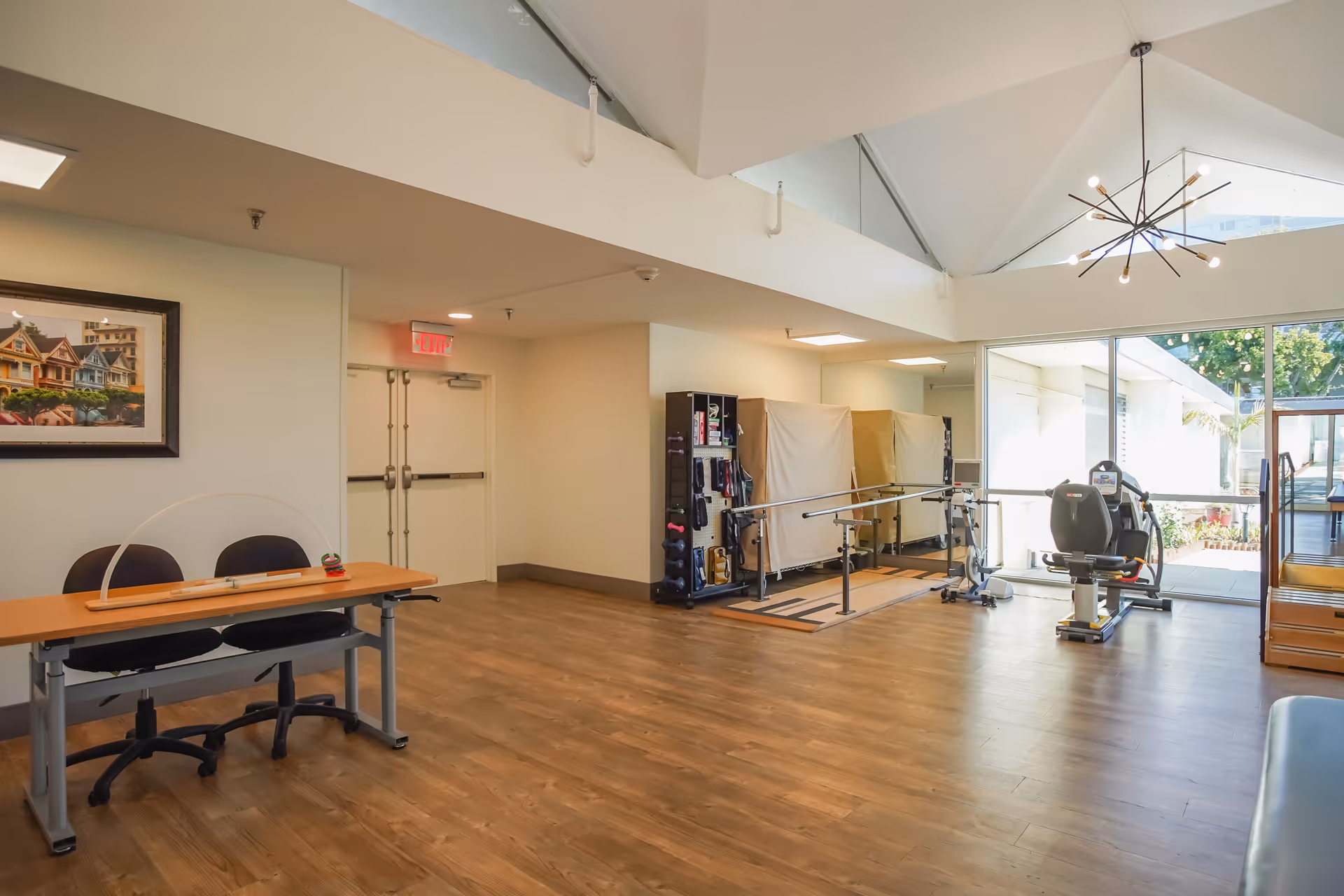 A spacious room with wooden flooring and a high ceiling featuring a modern chandelier. The room includes a table with two chairs and a clear protective barrier, a framed picture on the wall, and exercise equipment including parallel bars and a stationary bike near large windows that let in natural light. There is an exit door and a rack with various exercise tools.