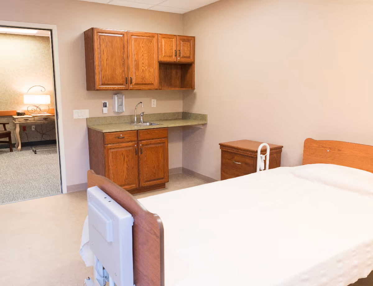 A simple senior bedroom with a single bed, wooden nightstand and built-in sink and cabinets against a beige wall.
