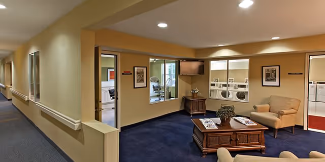 Interior view of a senior living facility hallway and common area at Hampshire Village. The hallway has beige walls with handrails and blue carpeting. The common area features a wooden coffee table with magazines, two armchairs, a wall-mounted TV, and windows looking into a laundry room with washers and dryers.