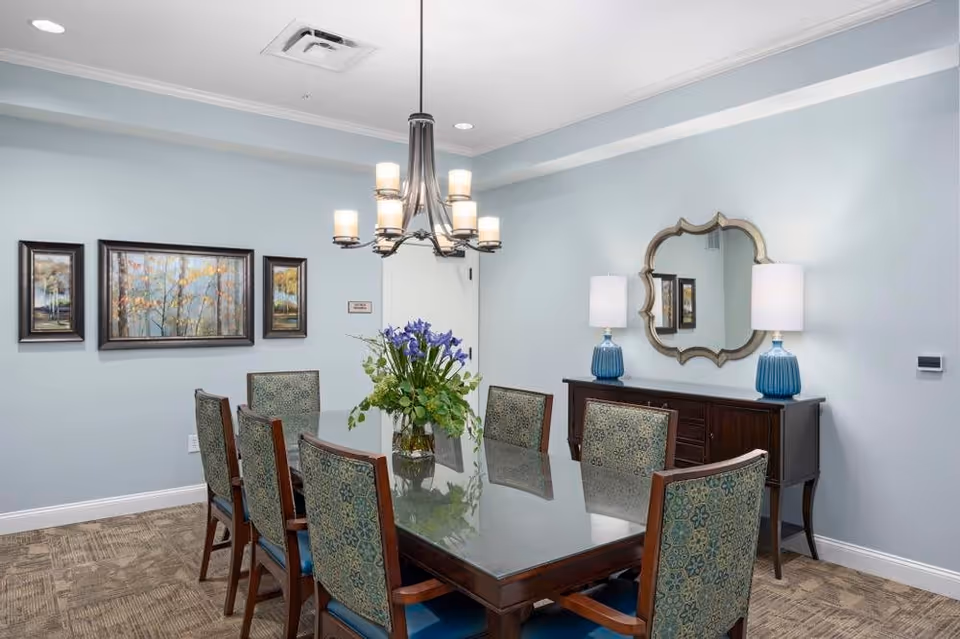 A dining room with a rectangular glass-top table surrounded by eight upholstered chairs with patterned backs. A vase with purple and green flowers is centered on the table. On the wall behind the table, there are three framed landscape paintings. A dark wooden sideboard with two blue lamps and a decorative mirror above it is against the right wall. The walls are painted light blue, and a modern chandelier with multiple lights hangs above the table.
