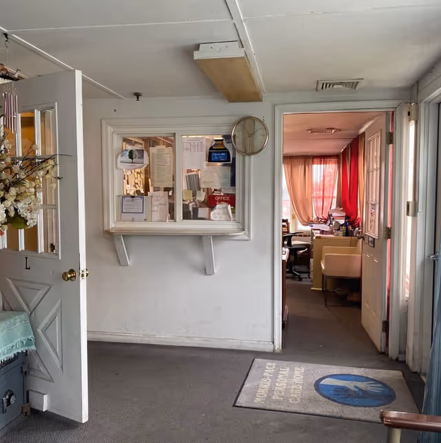 Interior view of a personal care home reception area with an open door on the left adorned with a floral wreath. A small office window with various papers and notices is mounted on the wall, with a clock above it. Through an open doorway, an office space with desks, chairs, and red curtains on the windows is visible. A floor mat with the text 'Morris Pace Personal Care Home' and a logo is placed on the carpeted floor.