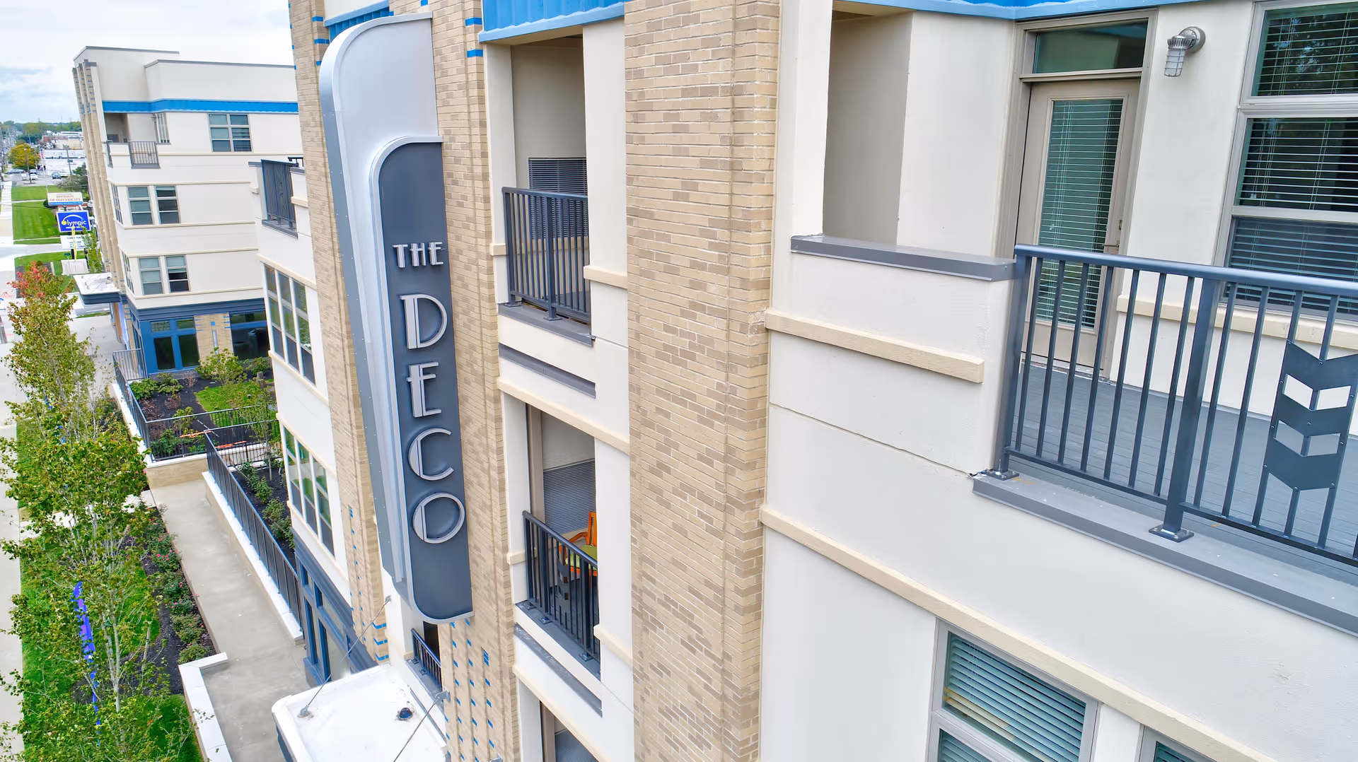 Exterior view of a multi-story building with beige brick and cream-colored walls, featuring balconies with black railings. A vertical sign on the building reads 'THE DECO'. Trees and a sidewalk are visible along the street beside the building.