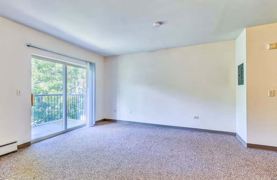 An empty living room with a sliding glass door to a balcony, neutral walls, and carpet.