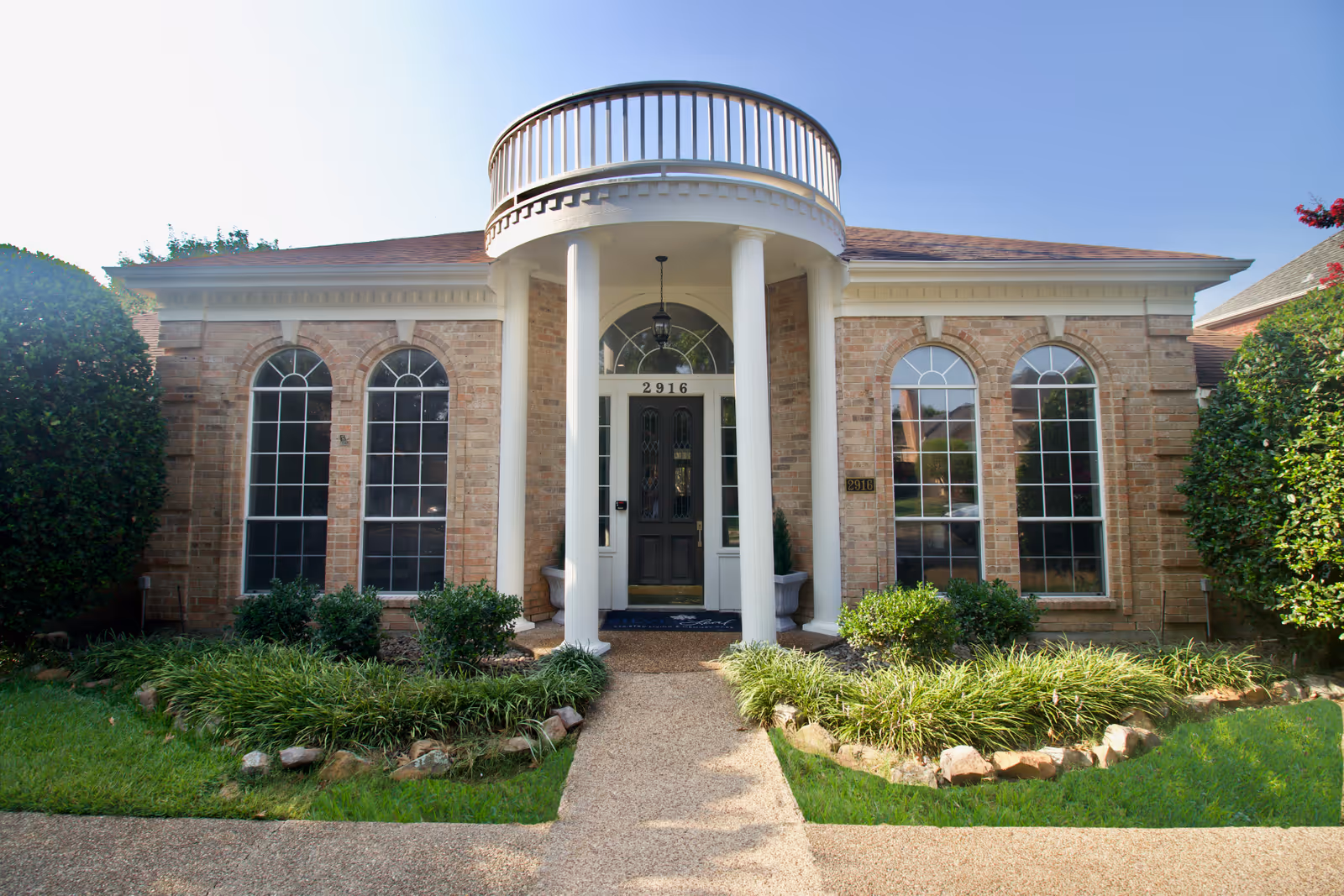 Front exterior view of a brick building with a central entrance featuring two white columns and a balcony above. The building has four tall arched windows, two on each side of the entrance, and is surrounded by green shrubs and grass.