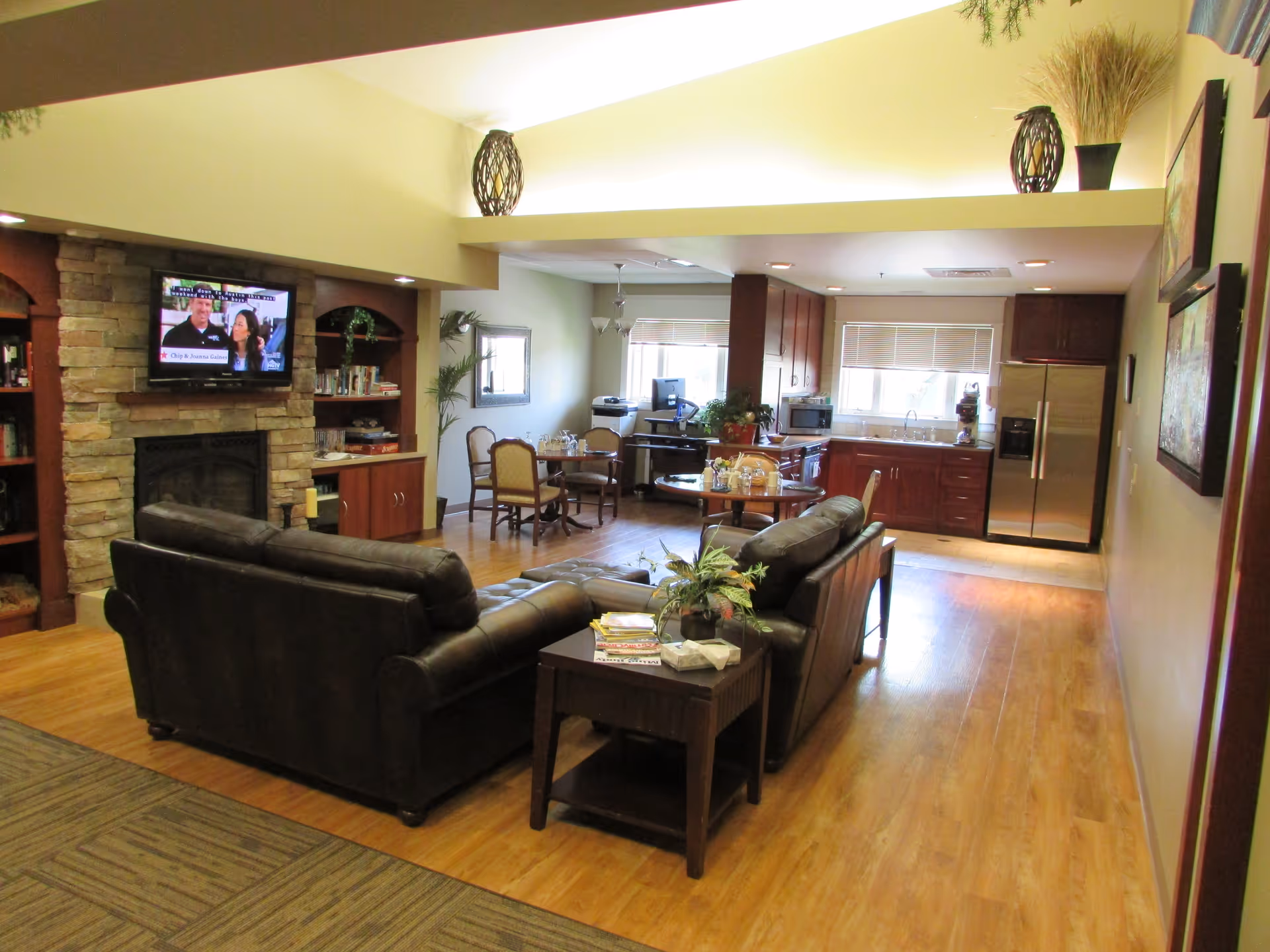 A cozy living area with two dark leather sofas arranged around a wooden coffee table with plants and books. In the background, there is a kitchen with wooden cabinets, a stainless steel refrigerator, and a dining area with a round table and chairs. A stone fireplace with a mounted flat-screen TV is on the left side of the room. The space has wooden flooring and warm lighting.