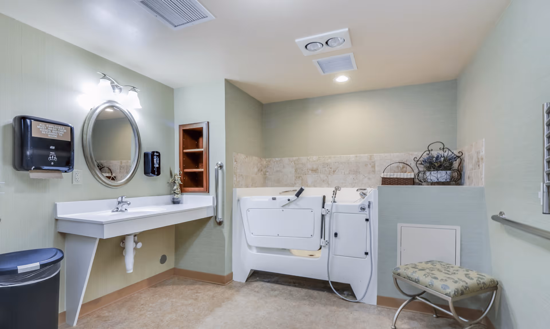 A clean and accessible bathroom featuring a walk-in bathtub with safety handles, a wall-mounted sink with a round mirror above it, a soap dispenser, a paper towel dispenser, a small cushioned bench, and a trash can. The walls are painted light green with beige tile accents around the bathtub area.