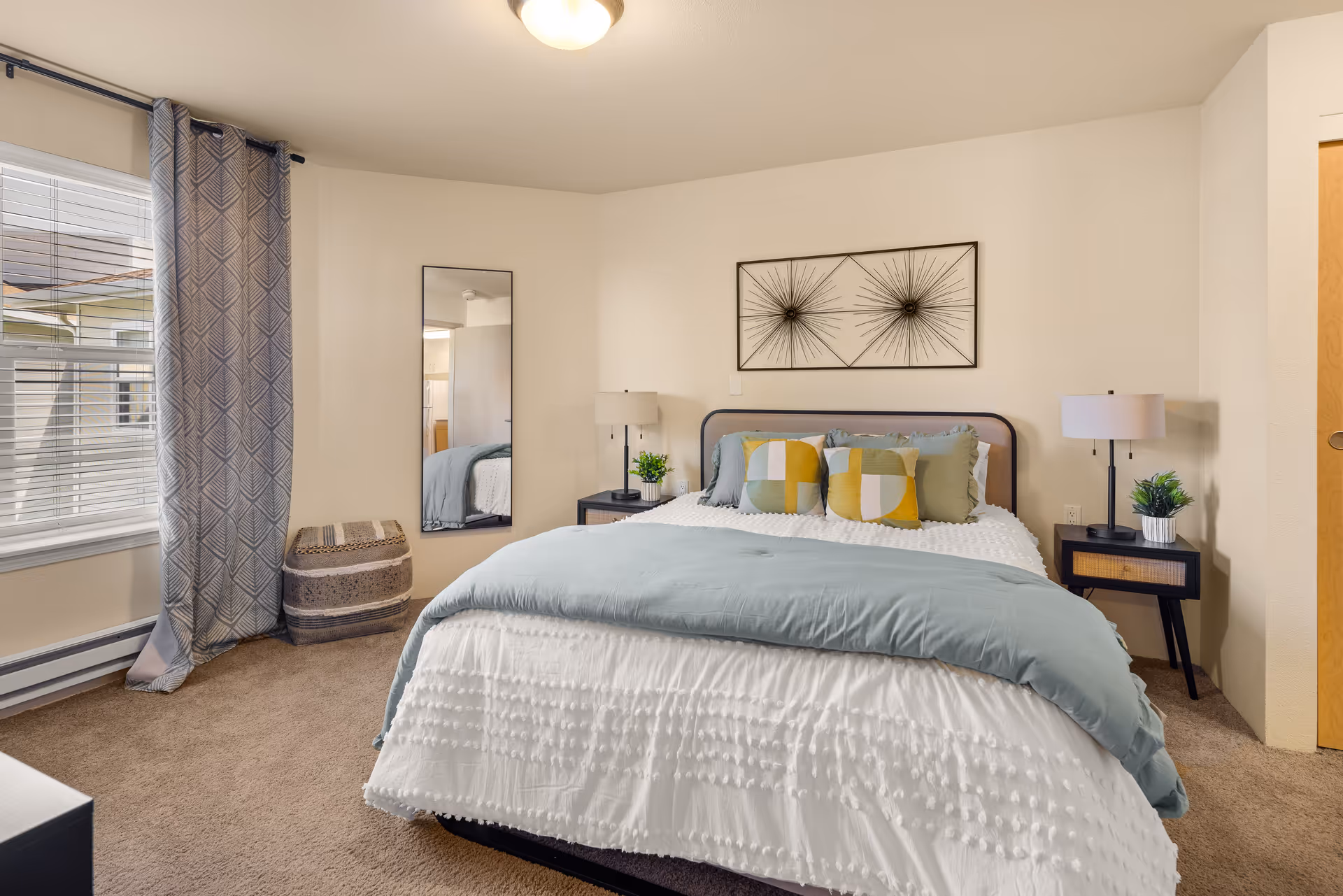 A cozy bedroom with a neatly made bed featuring a white textured comforter and a light blue blanket. The bed has four decorative pillows in shades of green, yellow, and white. On either side of the bed are black nightstands with table lamps and small potted plants. A large window with gray patterned curtains allows natural light into the room. A full-length mirror is mounted on the wall next to a woven basket. The walls are painted a light beige color, and the floor is carpeted in a neutral tone.