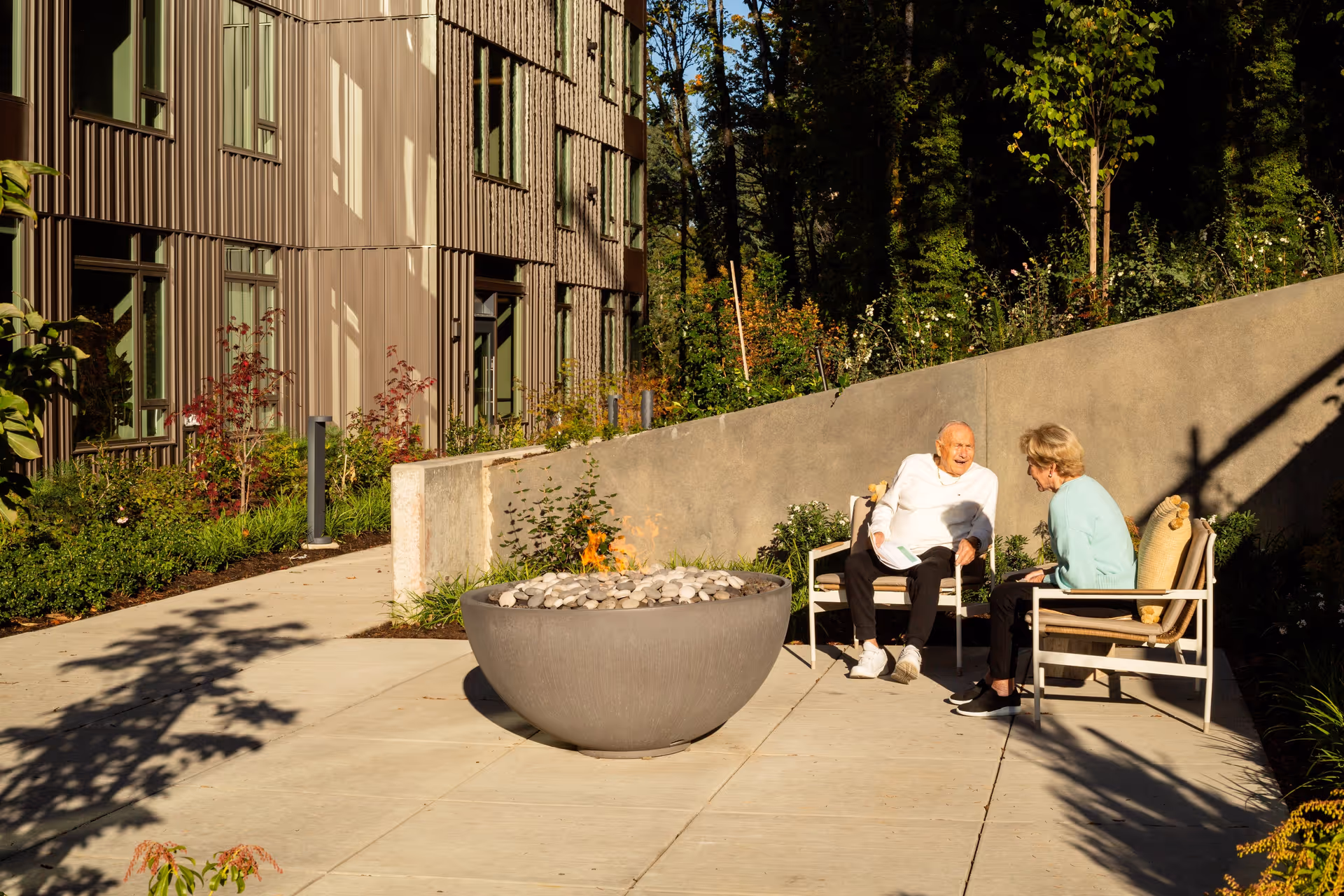 Two elderly people sitting on outdoor chairs around a modern fire pit in a sunny patio area next to a building with large windows and surrounded by greenery.
