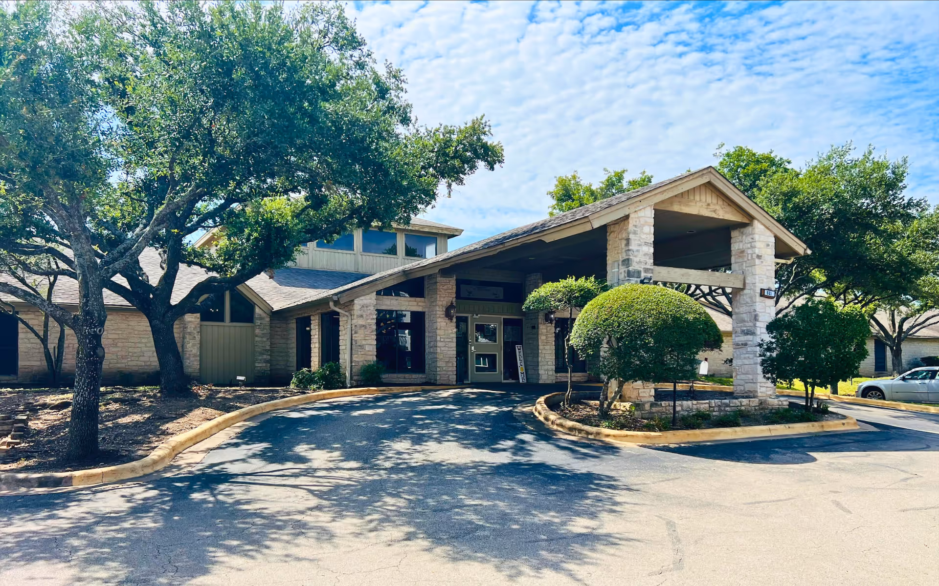 Front entrance and porte-cochere of a stone-faced senior living facility with driveway, trees, and landscaped shrubs under a partly cloudy sky.