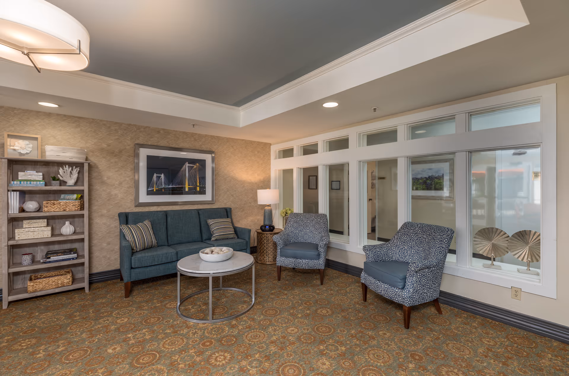 A cozy lounge with a teal sofa, two patterned armchairs, a round coffee table, bookshelf, and framed art on the wall.