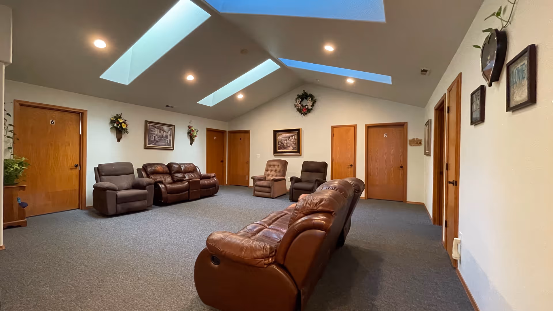 Communal lounge with leather recliners and sofas under skylights and recessed lighting, with multiple wooden doors along the walls.