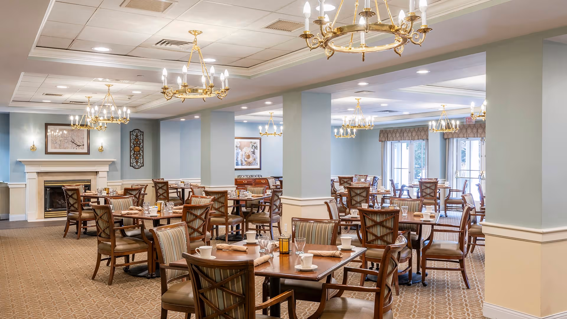 A spacious dining room with multiple wooden tables and chairs arranged neatly. Each table is set with cups, glasses, and napkins. The room features light blue walls, beige carpeting with a geometric pattern, and several elegant gold chandeliers hanging from the ceiling. There is a fireplace on one side with framed artwork above it, and large windows allowing natural light to enter the space.