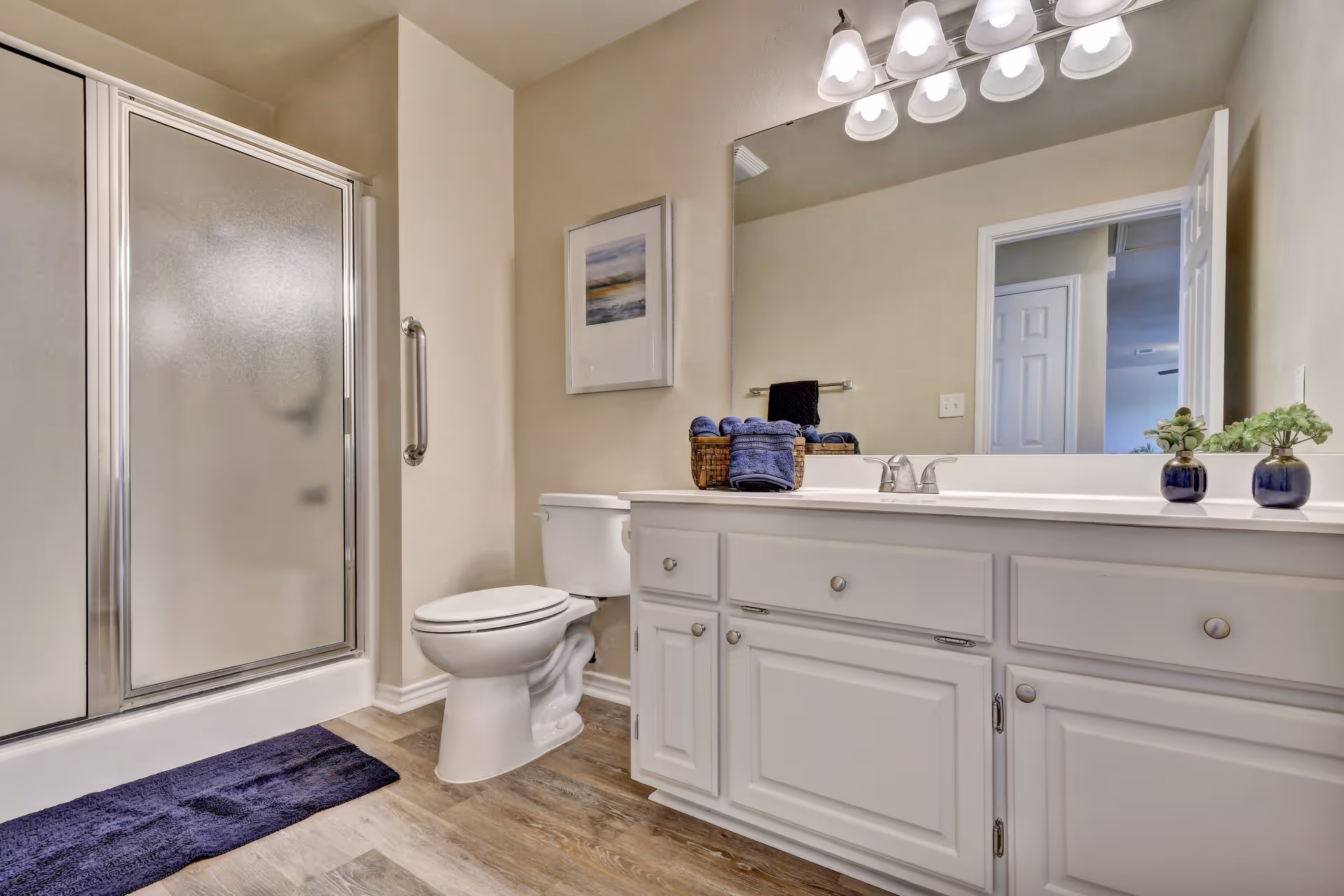 A clean and modern bathroom featuring a glass-enclosed shower with a silver frame, a white toilet, and a white vanity with multiple drawers and cabinets. On the vanity countertop are two small potted plants and a basket with rolled blue towels. Above the vanity is a large mirror with a row of five light fixtures. The floor has wood-like tiles, and a dark blue bath mat is placed in front of the shower. A framed picture hangs on the wall above the toilet.