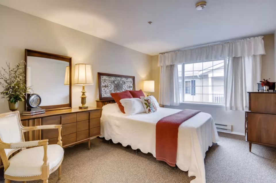 A cozy bedroom with a neatly made bed featuring white bedding and red accent pillows and throw. The room includes a wooden dresser with a large mirror, a table lamp, a wooden chair with white upholstery, a window with white curtains, and a small wooden chest of drawers with decorative items on top.