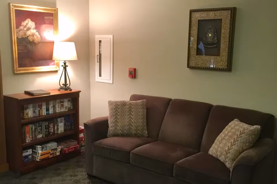 A cozy seating area with a brown sofa and patterned pillows, a bookshelf of games, a lamp, and framed artwork on the walls.