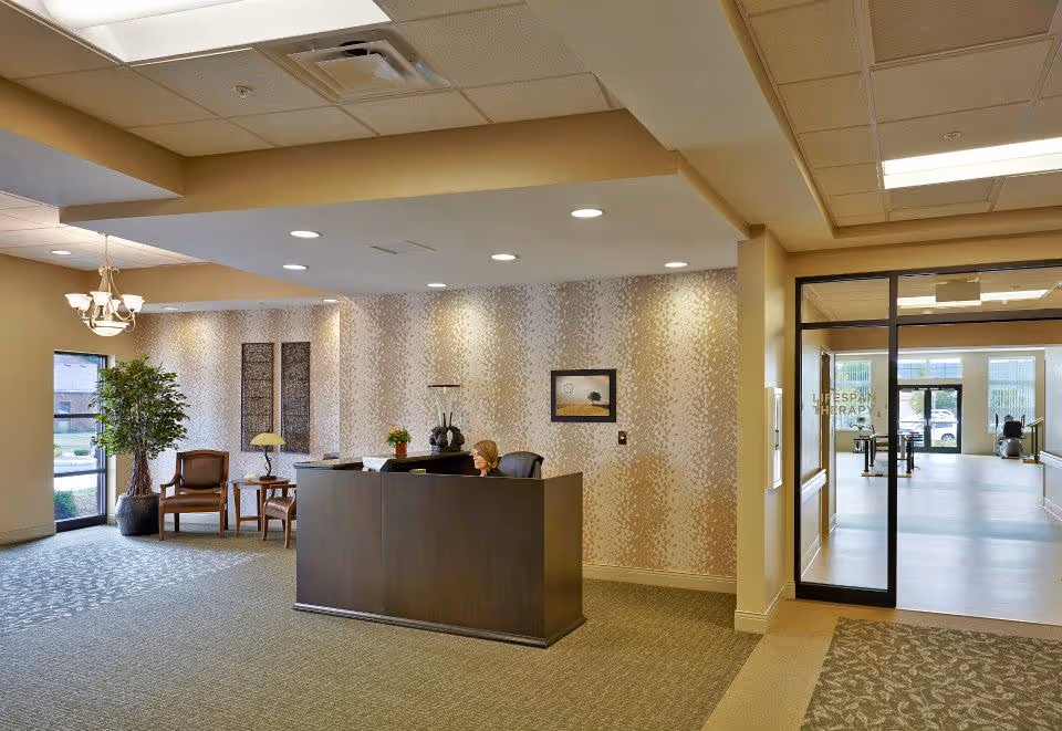 Reception area of Harbour Manor & The Lodge with a dark wood front desk, a receptionist seated behind it, patterned wallpaper, two wooden chairs with a small table and lamp, a large potted plant near a window, and a glass door leading to a therapy room with exercise equipment.