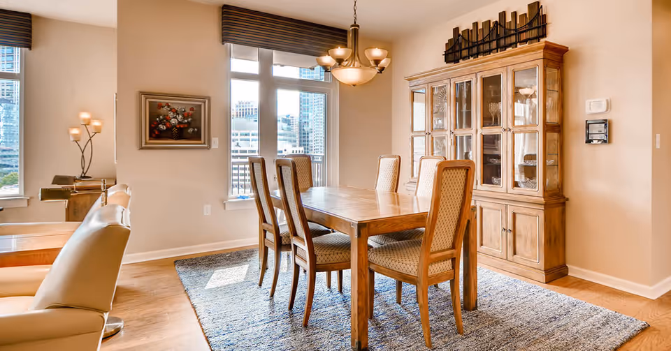 Sunlit dining room with a wooden table and six chairs, a glass-front china cabinet, chandelier, and large windows showing a city view.
