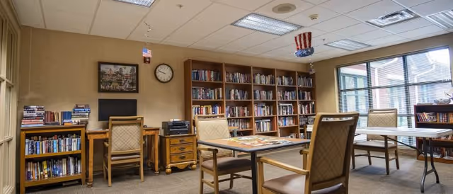 A well-lit room with bookshelves filled with books along the walls, several tables with chairs arranged around them, a desk with a computer and printer, and patriotic decorations hanging from the ceiling. Large windows allow natural light to enter the room.