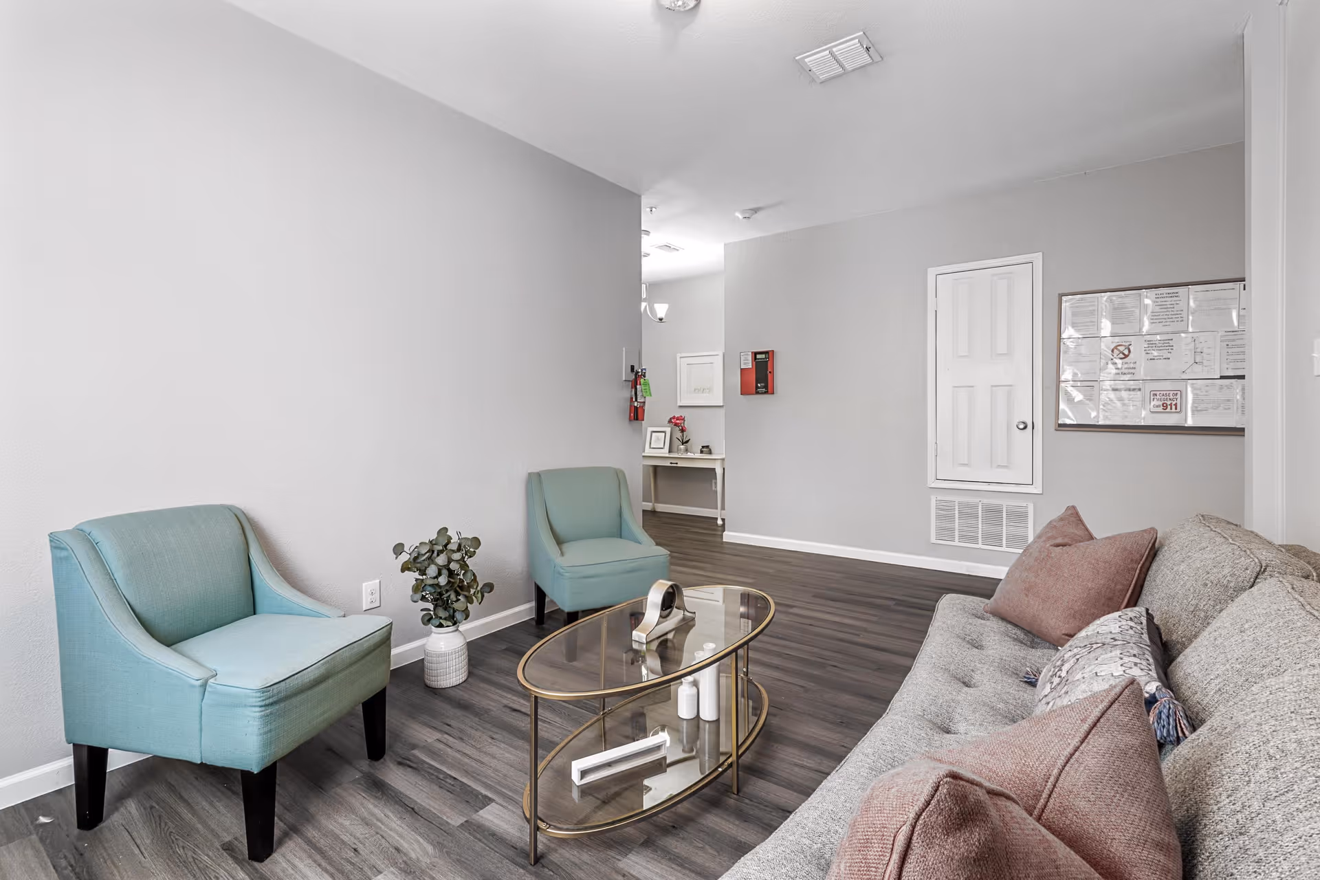 A modern senior living facility common area with two teal armchairs, a glass coffee table with decorative items, and a gray sofa with pink and patterned pillows. The room has light gray walls, dark wood flooring, and a small table with framed pictures and flowers in the background.