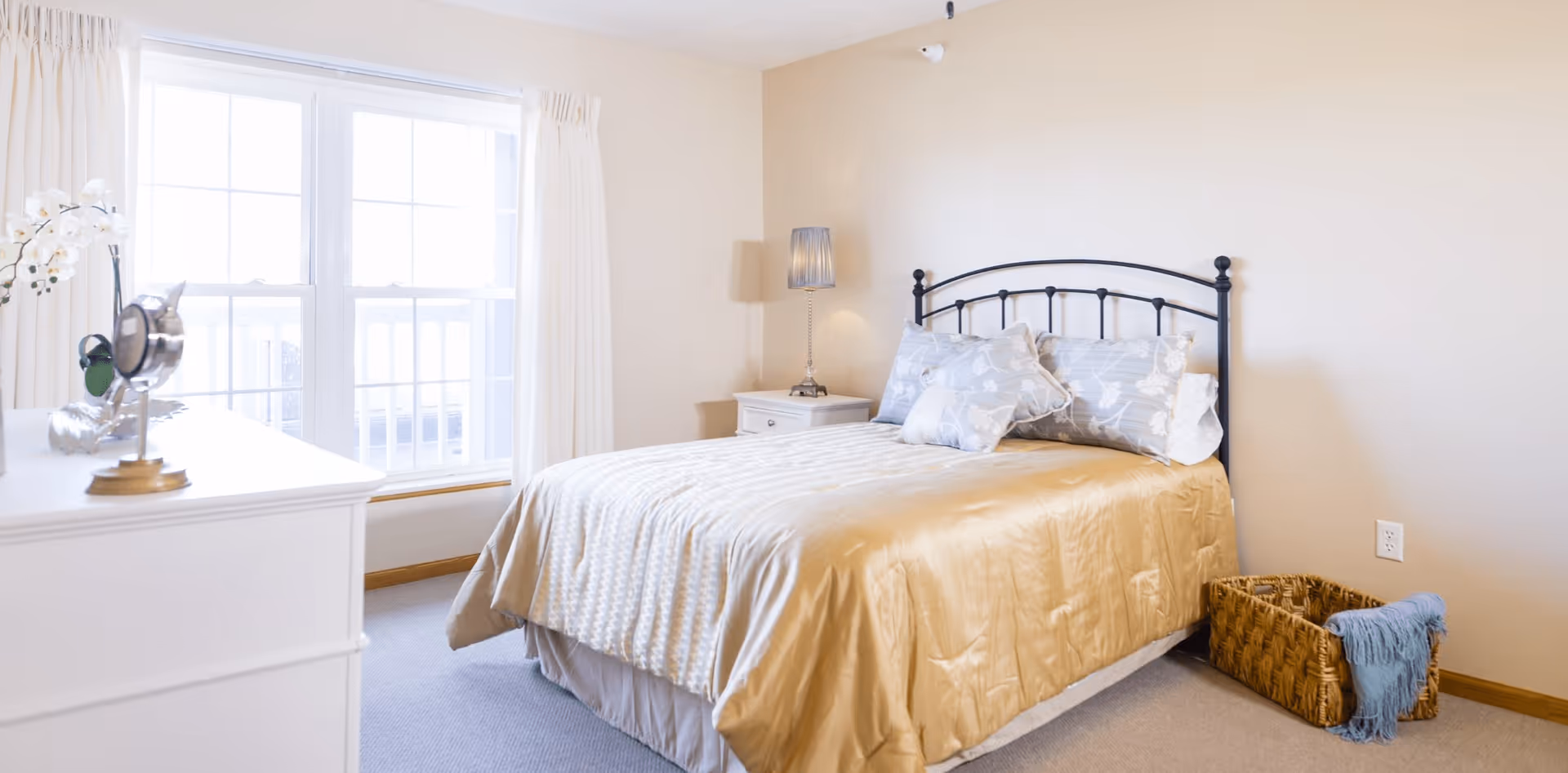A bright bedroom with a large window covered by white curtains, a bed with a black metal headboard, beige bedding, and decorative pillows. There is a white dresser with a mirror and decorative items on top, a nightstand with a lamp, and a woven basket with a blue blanket on the floor.
