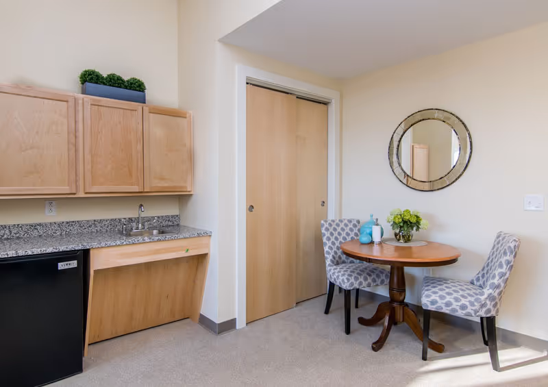 Small kitchenette with a granite countertop and cabinets beside a round dining table with two patterned chairs and a wall mirror.
