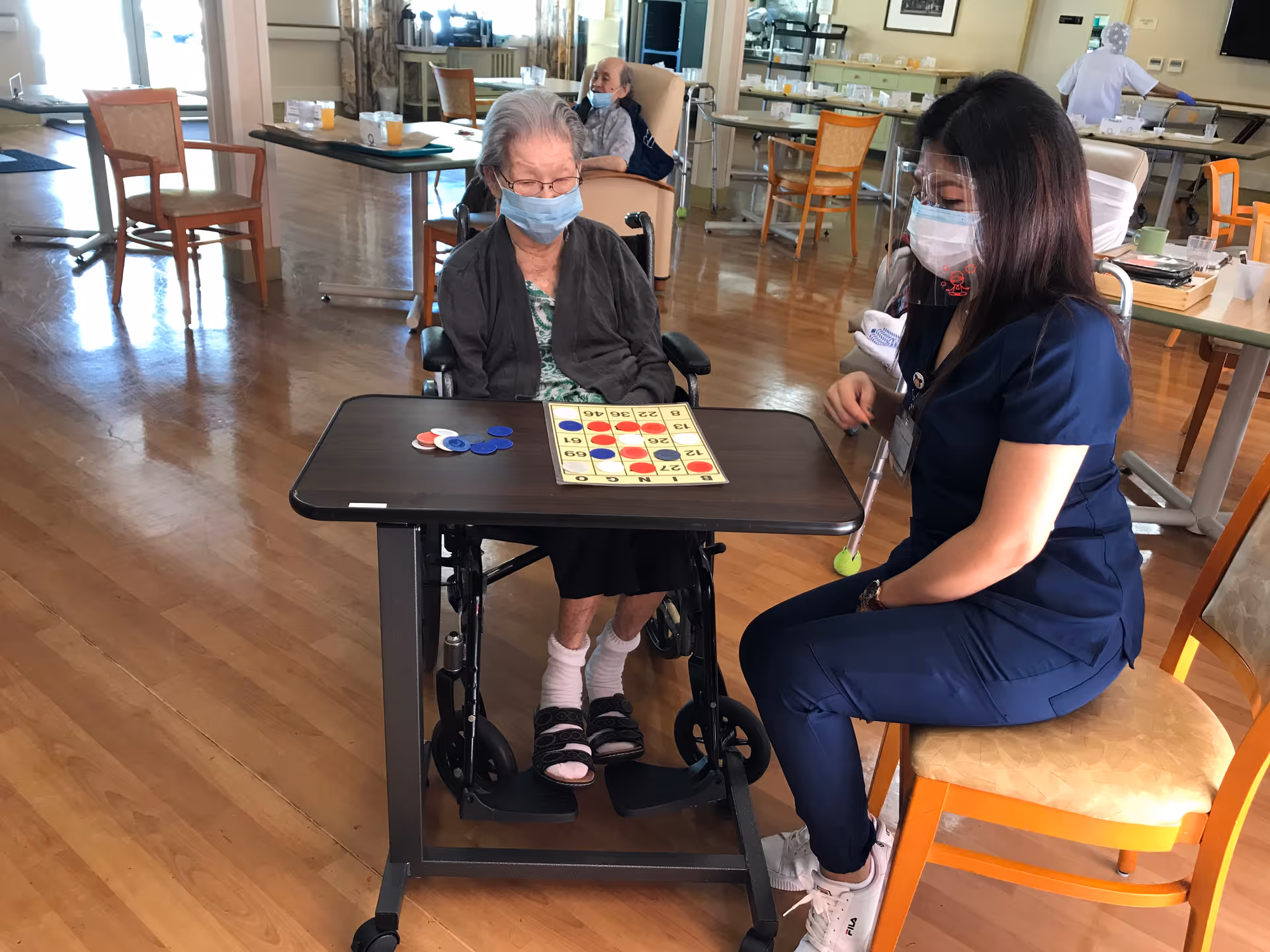 An elderly woman in a wheelchair wearing a mask is playing a game with a caregiver who is also wearing a mask and face shield. They are seated at a table in a spacious room with wooden floors and several chairs and tables in the background.