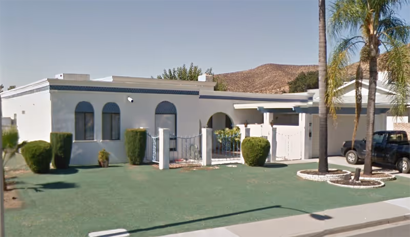 Exterior view of Sandy Lodge Care Home: white single-story building with arched windows and palm trees.