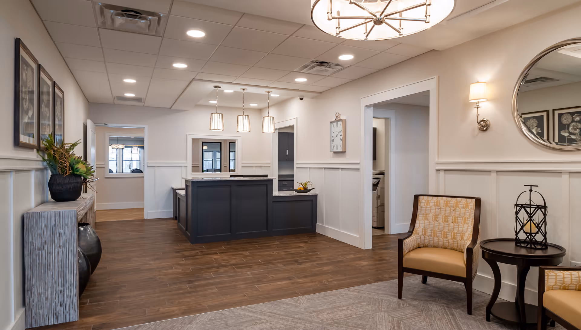A modern and clean reception area in a senior living facility with a dark gray reception desk, three pendant lights hanging above it, wood flooring, and beige walls with white wainscoting. There are two patterned armchairs with a small round table between them on the right side, a large round mirror and a wall sconce above them. On the left side, there is a console table with decorative plants and framed artwork on the wall. The ceiling has recessed lighting and a large chandelier.