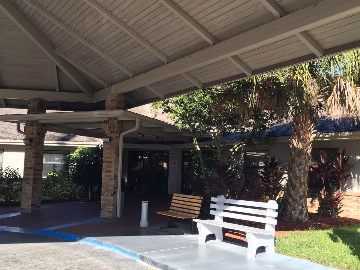 Covered entrance area of a building with brick pillars, a white bench, a wooden bench, and landscaping including palm trees and bushes.