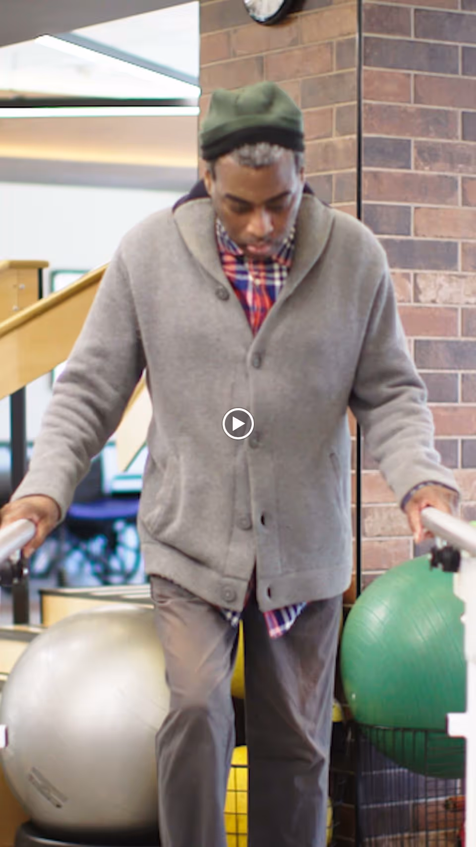 An elderly man wearing a gray cardigan and green hat is using parallel bars for support while walking in a rehabilitation facility. Behind him are large exercise balls and a brick wall.