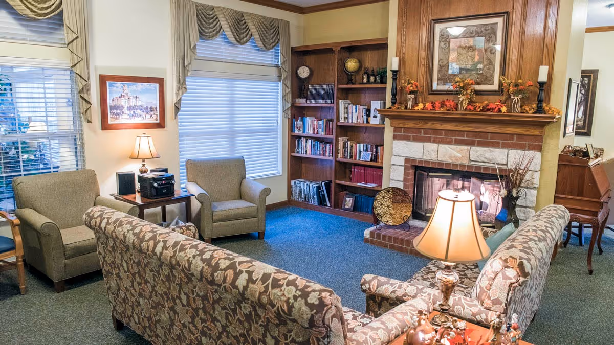 A cozy living room with floral patterned sofas, two armchairs, a wooden bookshelf filled with books, a fireplace decorated with autumn-themed ornaments, a table lamp, and framed artwork on the walls. The room has large windows with blinds and valances, and a piano is visible in the background.