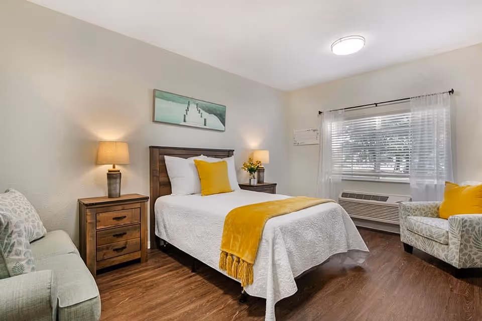 A cozy bedroom with a wooden bed frame, white bedding, and yellow accent pillows and throw blanket. There are two wooden nightstands with lamps on either side of the bed. A green patterned armchair with a yellow pillow is placed near a window with white curtains and blinds. The floor is wooden, and a painting of a pier over water hangs above the bed.