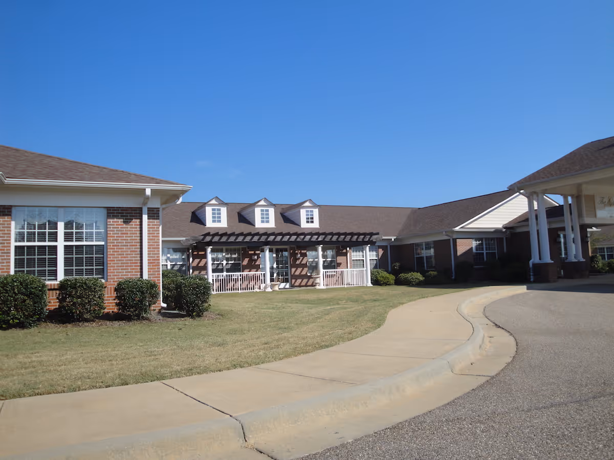Exterior view of The Gables at Charlton Place Assisted Living facility showing a brick building with white-trimmed windows, a covered entrance with white columns, a pergola over a porch area, and a curved driveway under a clear blue sky.
