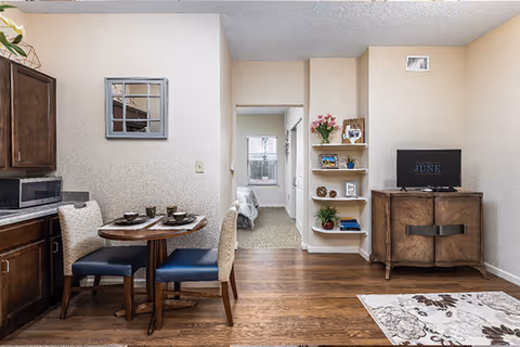 Open-plan living area with a small dining table and chairs, kitchenette cabinets, a TV console, built-in shelves, and a view into a bedroom.